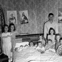 Woman in bed holding 2 infants, surrounded by her husband and children